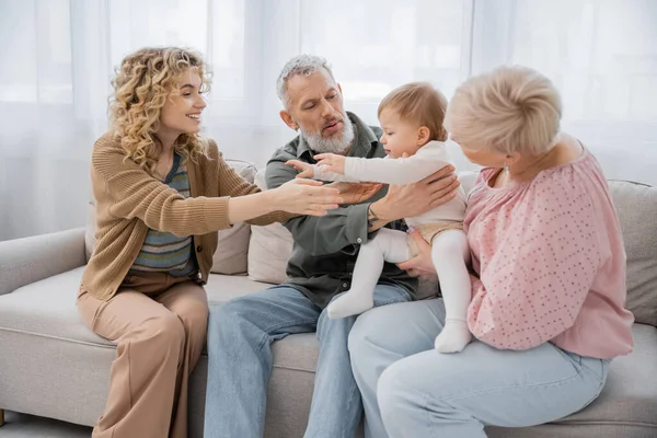 Cheerful blonde woman outstretching hands to little daughter near parents on couch in living room — Stock Photo