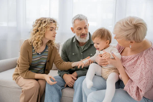 Curly woman calming upset baby near mature parents on sofa in living room — Stock Photo
