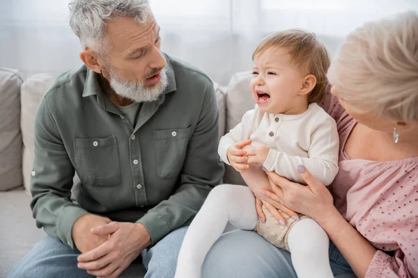 Confused middle aged couple calming crying granddaughter on couch in living room — Stock Photo