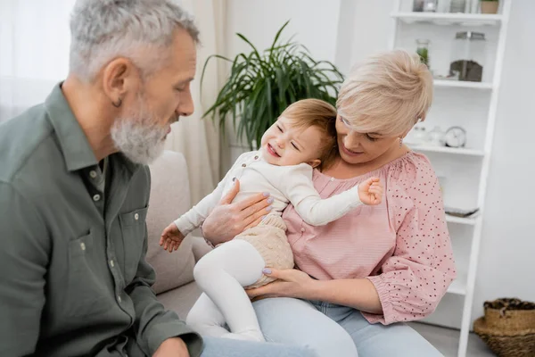 Reife Frau hält unzufriedene Enkelin, während sie neben dem bärtigen Ehemann im Wohnzimmer sitzt — Stockfoto