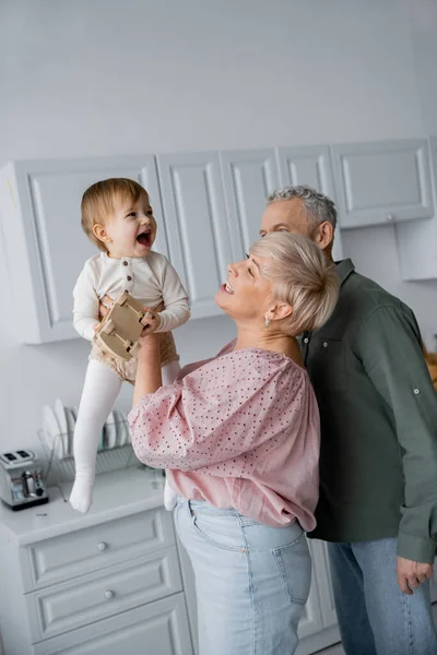 Chica excitada con coche de juguete riendo cerca de abuelos felices en la cocina - foto de stock