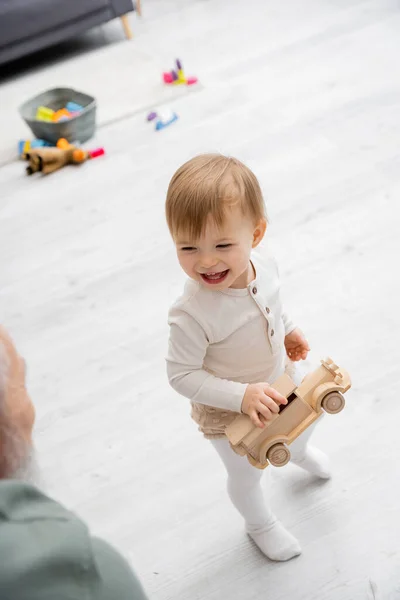 Visão de alto ângulo da criança feliz de pé com carro de brinquedo perto do avô borrado na sala de estar — Fotografia de Stock