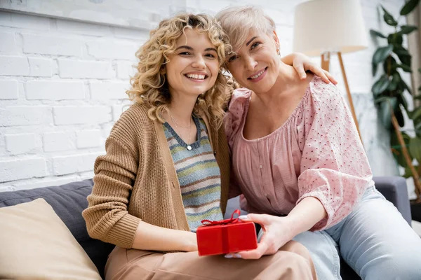 Femme blonde aux cheveux ondulés embrassant mère heureuse tenant boîte cadeau tout en étant assis sur le canapé à la maison — Photo de stock