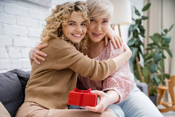 Happy mature woman with adult daughter embracing and looking at camera near gift box in living room — Stock Photo