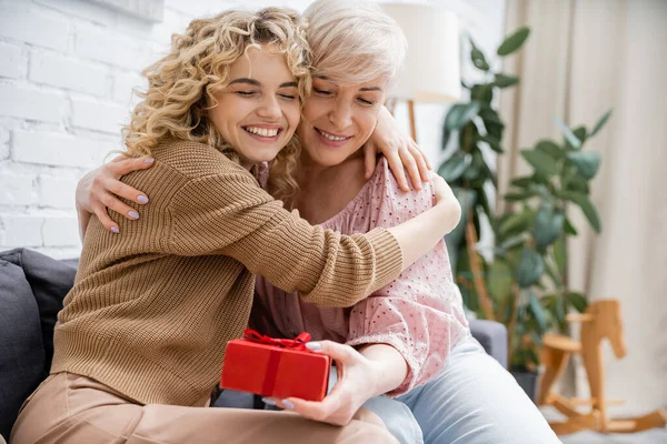 Overjoyed mature woman with closed eyes holding present and embracing with happy daughter in living room — Stock Photo