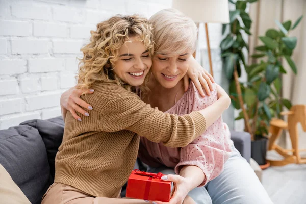 Joyful woman with closed eyes embracing happy middle aged mother sitting with gift box on couch in living room — Stock Photo