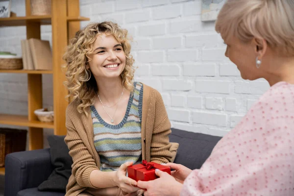 Mujer alegre con cabello rubio ondulado presentando regalo a la madre mientras está sentada en el sofá en la sala de estar - foto de stock