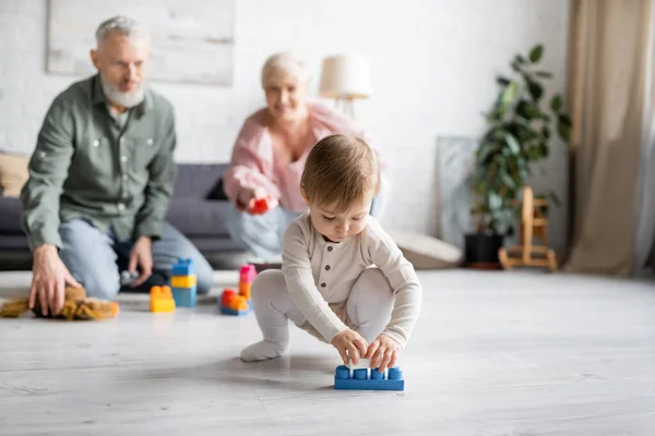Baby girl playing with building blocks on floor in living room near grandparents on blurred background — Stock Photo