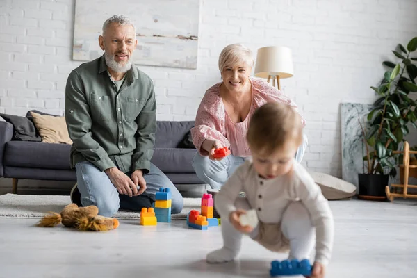 Happy middle aged grandparents sitting on floor near building blocks while little girl playing with toy car in living room — Stock Photo