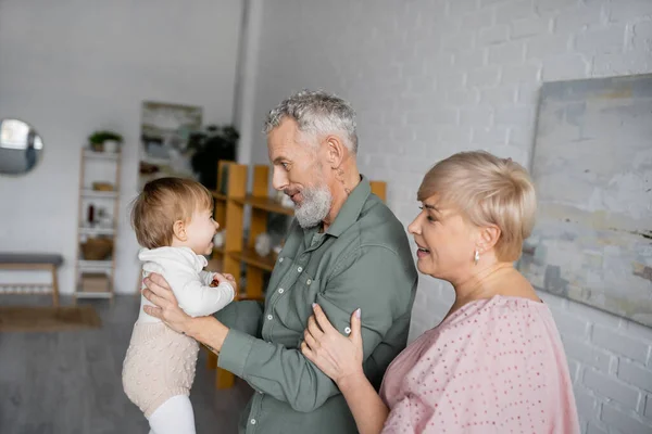 Вид сбоку босоногого мужчины, держащего плачущую внучку рядом с улыбающейся женой в гостиной — стоковое фото