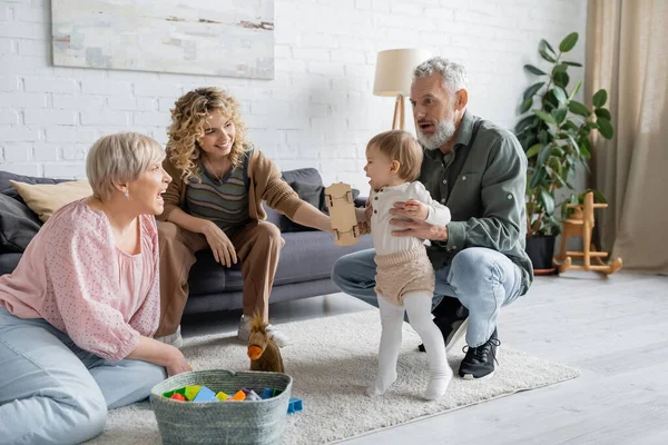 Cheerful bearded man supporting baby girl doing her first steps near family in living room — Stock Photo