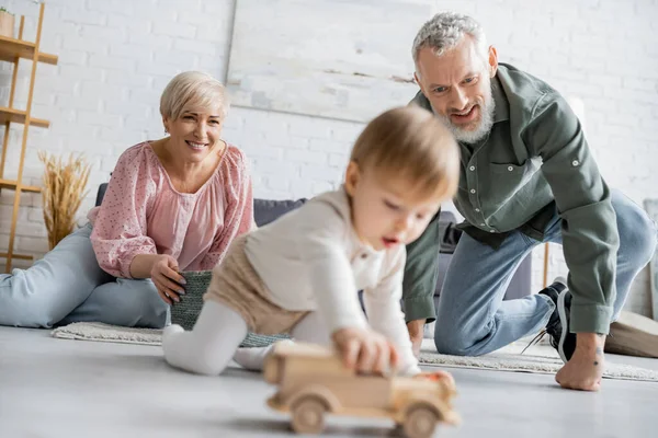 Pareja de mediana edad llena de alegría mirando borrosa nieta jugando con coche de juguete en el suelo en la sala de estar - foto de stock