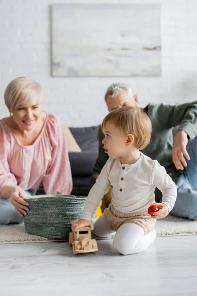 Little child playing with toy car near wicker basket and smiling grandparents on blurred background in living room — Stock Photo