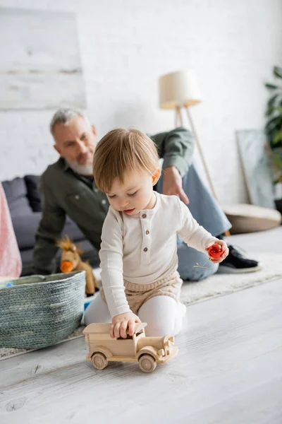 Little girl playing with toy car on floor in living room near grandpa on blurred background — Stock Photo