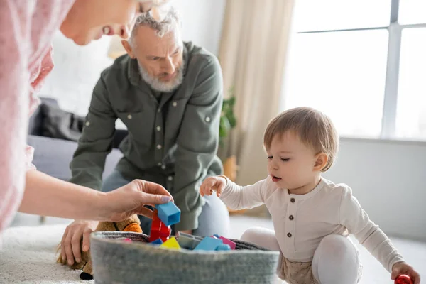 Little girl looking at toys in wicker basket while playing with grandparents in living room — Stock Photo