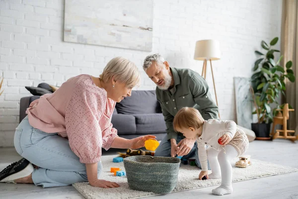 Pareja de mediana edad jugando con la nieta del niño en la alfombra de piso en la sala de estar moderna - foto de stock
