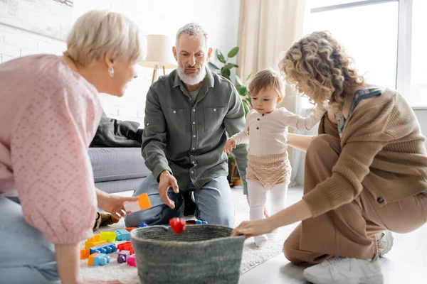 Blonde woman with toddler daughter and mature parents near wicker basket and toys on floor in living room — Stock Photo