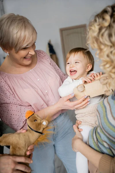 Bambino spensierato che tiene macchina giocattolo e ride vicino ai nonni e alla madre in soggiorno — Foto stock
