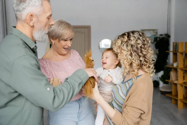 Bärtiger Mann hält Spielzeugpferd neben fröhlicher Enkelin und Familie im Wohnzimmer — Stockfoto