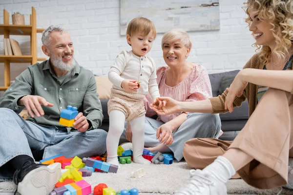 Femme joyeuse tenant la main de la petite fille près des parents heureux et des jouets sur le sol dans le salon — Photo de stock