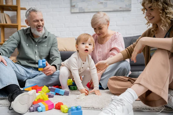 Niña mirando lejos cerca de los juguetes y la familia feliz en el suelo en la sala de estar - foto de stock