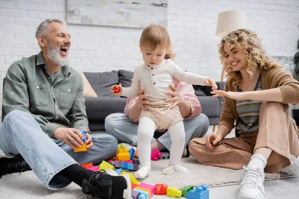 Hombre barbudo emocionado riendo cerca de la familia jugando con el niño en el suelo en la sala de estar - foto de stock