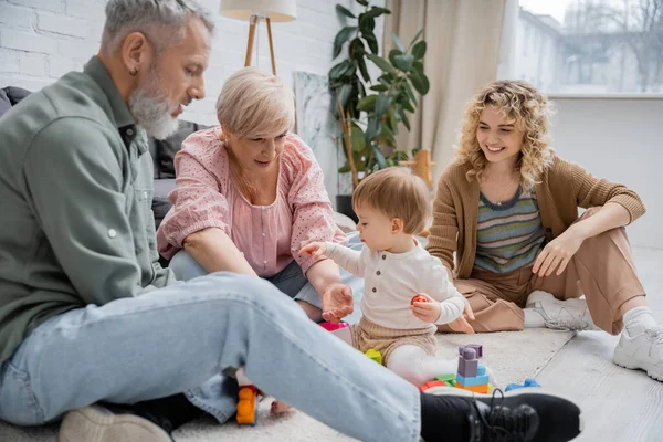 Mulher alegre olhando para os pais de meia-idade jogando blocos de construção jogo com criança no chão na sala de estar — Fotografia de Stock