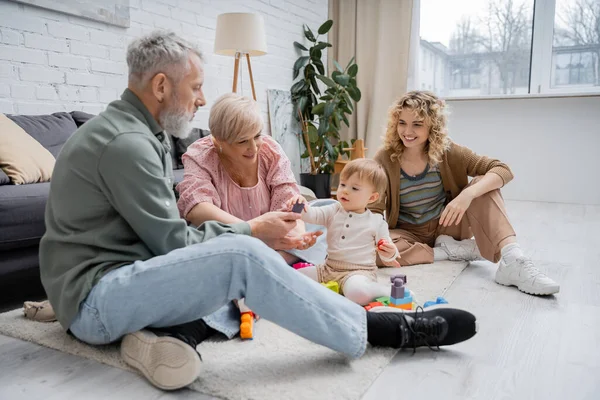 Fröhliches reifes Paar spielt Bauklötzchenspiel mit Enkelin, während es im modernen Wohnzimmer auf dem Boden sitzt — Stockfoto