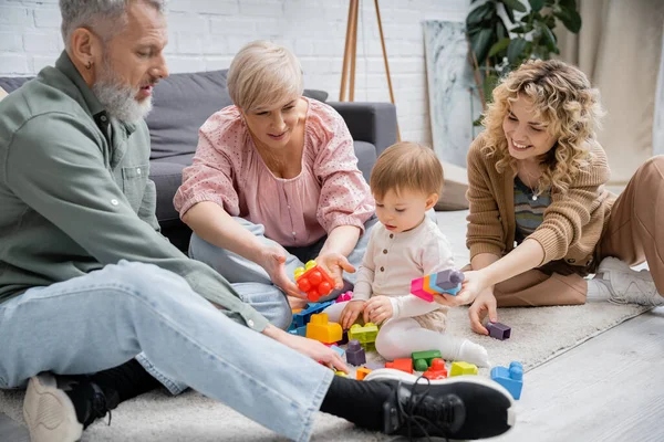 Bambina che gioca con blocchi di costruzione colorati vicino a madre e nonni felici sul pavimento in soggiorno — Foto stock