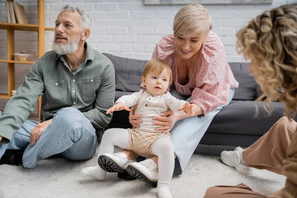 Happy middle aged woman playing with little granddaughter near family in living room — Stock Photo
