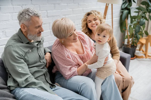 Baby girl looking at camera near cheerful mother and grandparents on couch in living room — Stock Photo