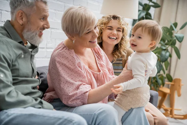 Toddler girl smiling near cheerful grandparents and mother on couch in living room — Stock Photo