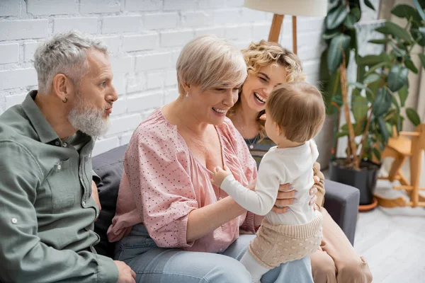 Mujer llena de alegría sosteniendo pequeña nieta cerca de la familia feliz en el sofá en casa - foto de stock