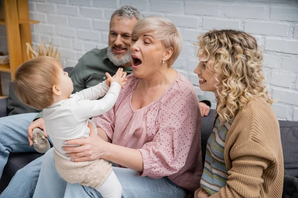Grandmother opening mouth while having fun with toddler girl on sofa in living room — Stock Photo