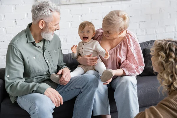 Criança despreocupada rindo perto de avós felizes e mãe turva na sala de estar — Fotografia de Stock