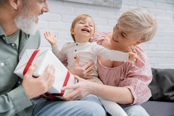 Joyeux geste d'enfant et souriant avec les yeux fermés près des grands-parents avec présent dans le salon — Photo de stock