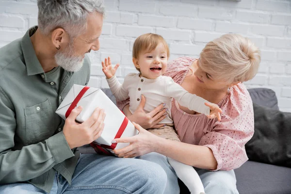 Criança feliz gestos perto de avós e caixa de presente na sala de estar — Fotografia de Stock