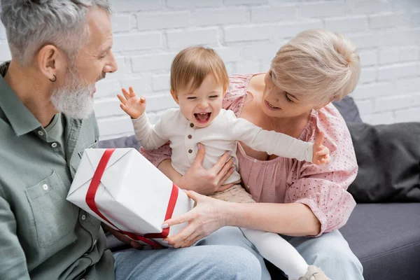 Carefree baby laughing and gesturing near smiling grandparents and present on couch at home — Stock Photo