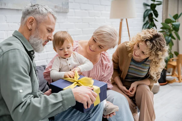 Mature bearded man holding present near toddler granddaughter and smiling family in living room — Stock Photo