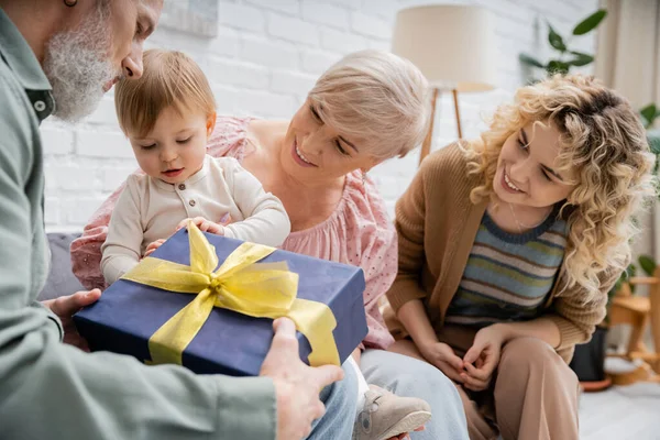 Bärtiger Mann hält Geschenkbox in der Nähe von Enkelin und Familie im Wohnzimmer — Stockfoto