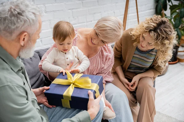 Niño pequeño abrir caja de regalo cerca de la madre sonriente y abuelos en el sofá en la sala de estar - foto de stock