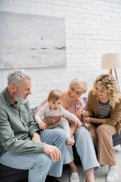 Toddler girl looking at camera near mother and grandparents on sofa in living room — Stock Photo