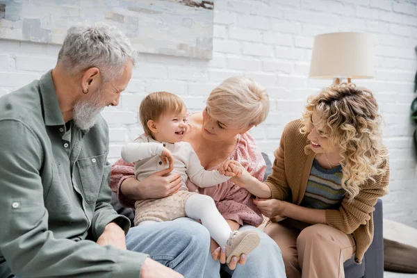 Взволнованная девочка смеется рядом с улыбающейся матерью и бабушкой и дедушкой, сидя на диване в гостиной — стоковое фото