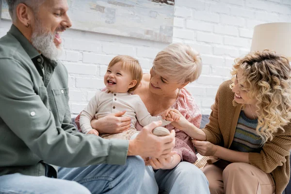 Bearded man touching leg of overjoyed granddaughter while having fun with family on couch in living room — Stock Photo