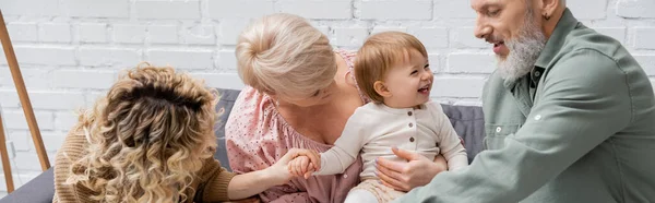 Счастливая женщина средних лет обнимает смеющуюся внучку рядом с семьей в гостиной, баннер — стоковое фото
