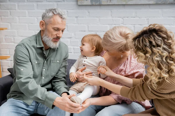 Mature couple touching leg of cheerful granddaughter while sitting on sofa in living room — Stock Photo