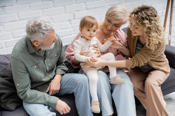 Улыбающаяся женщина держит за руки малышку дочь, проводя время с родителями на диване дома — стоковое фото
