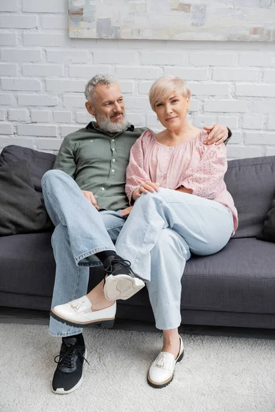 Happy bearded man embracing middle aged wife while sitting on couch at home — Stock Photo