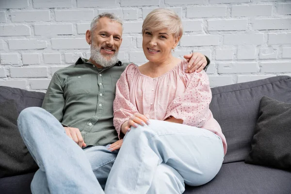 Joyful middle aged couple sitting on sofa in living room and smiling at camera — Stock Photo