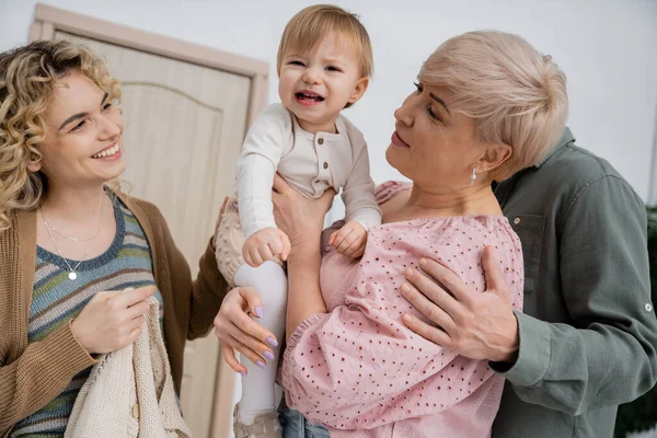 Cheerful woman looking at toddler daughter smiling in hands of grandmother in hall of apartment — Stock Photo
